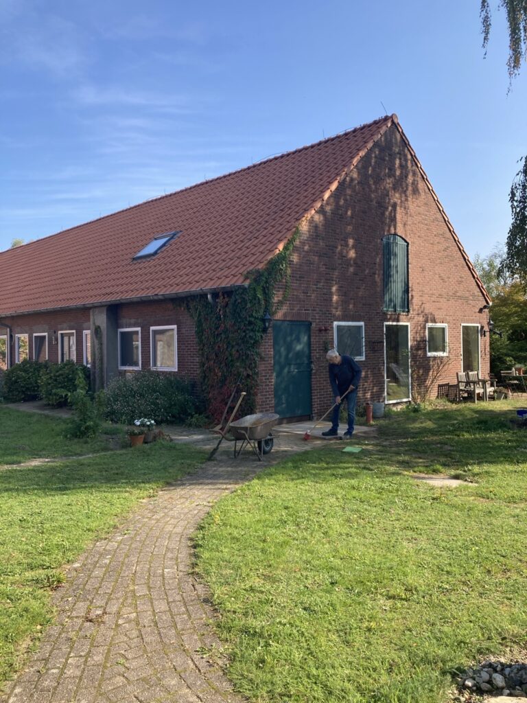 Alex veegt het laatste stukje nieuwe straatwerk bij de warmtepomp, met kruiwagen naast zich, voor een rode baksteenvleugel van de Leonardushoeve